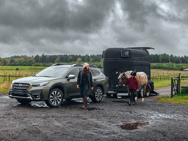 Subaru Outback purve su arklio priekaba, šalia eina moteris ir mergaitė – mergaitė veda arklį.